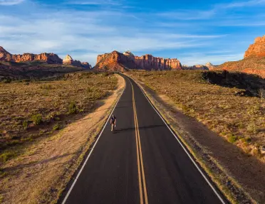 A lone cyclist rides on a paved road through a desert landscape framed by Zion’s red rock mesas, cliffs, and expansive mountain vistas under streaked clouds.
