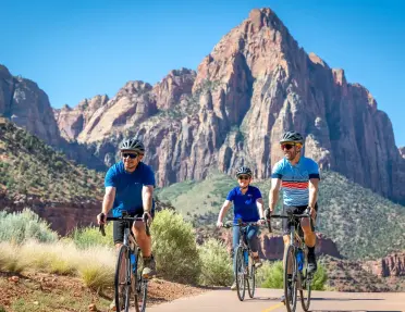 Three cyclists ride along a paved path beneath dramatic red rock cliffs and towering sandstone peaks in Zion National Park, surrounded by desert vegetation and clear blue skies.