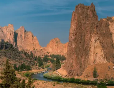 View of Smith Rock State Park featuring towering rock spires and cliffs rising above the Crooked River, which winds through a high desert canyon dotted with juniper trees.