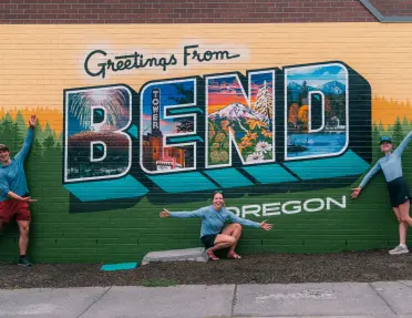Three people pose with outstretched arms in front of a colorful “Greetings From Bend, Oregon” mural depicting local landmarks, forests, mountains, and scenic landscapes.