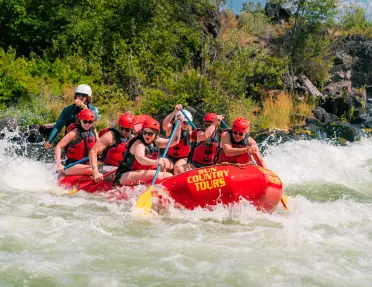 Group of rafters wearing helmets and life jackets paddle a red raft through whitewater rapids on a river near Smith Rock, surrounded by rocky banks and green vegetation.