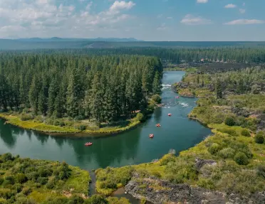 Aerial view of a winding river flowing through a landscape of pine forest, lava rock formations, and wetlands, with several rafting groups visible on the water.