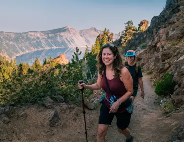 Two hikers walk along a rocky mountain trail overlooking a deep blue crater lake surrounded by rugged cliffs and evergreen trees. The scenic landscape is lit by soft natural light.