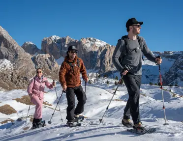 Two men and one woman smiling while hiking on a snowy valley, with hiking poles and snow shoes