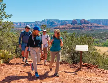 Group of hikers walking along a red dirt trail overlooking Sedona’s red rock formations and desert landscape under a clear blue sky.