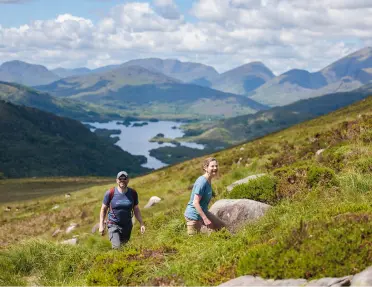 Two people ascending a grass and moss-covered hill, with a lake and mountains in the distance