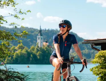 Cyclist pauses beside a turquoise lake with a church-topped island visible in the background. Forested hills and calm waters frame the scenic setting beneath a bright blue sky.