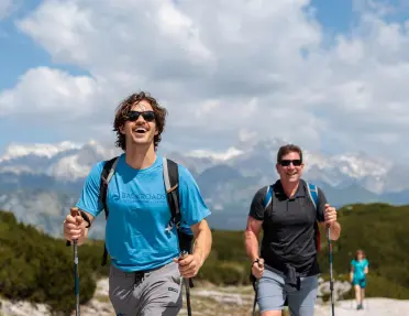 Hikers using trekking poles walk along a high alpine trail with snow-capped mountains stretching across the background. The rocky landscape and expansive mountain views are framed by a sky filled with scattered clouds.