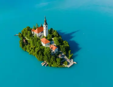 Aerial view of a small island covered in trees and historic buildings, centered around a church with a tall bell tower. The island sits in the middle of a vivid turquoise lake with boats docked along the shoreline.