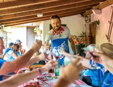 Cyclists gather around a long table at a rustic outdoor restaurant, raising glasses in a toast beneath a covered terrace with wooden beams and checkered tablecloths.