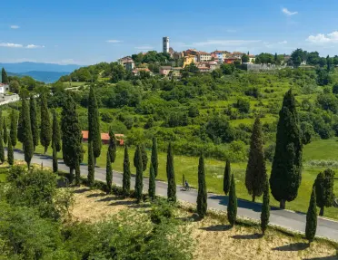 Aerial view of a cyclist riding along a road lined with tall cypress trees through rolling green countryside. A hilltop village with stone buildings and a church tower overlooks the landscape beneath a clear blue sky.