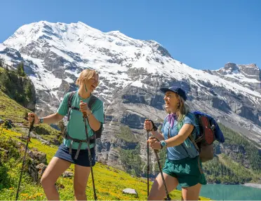 Two women looking at each other, smiling while hiking in front of a tall mountain