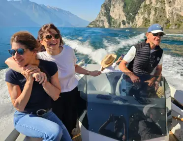 Passengers ride in a motorboat across a blue lake bordered by steep limestone cliffs and mountain scenery, with the boat's wake trailing behind on a clear day.