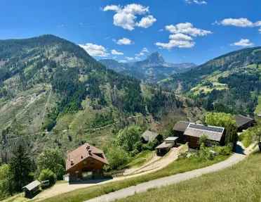 Panoramic view of a rural alpine valley featuring traditional farmhouses, winding roads, forested hillsides, and a distinctive mountain peak in the distance under a bright blue sky.