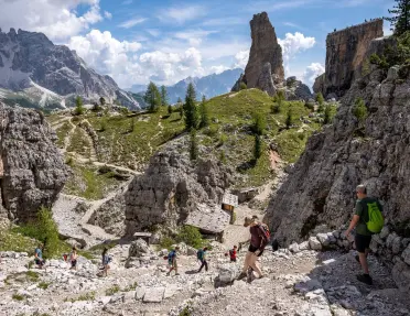 Hikers descend a rocky trail through a dramatic alpine landscape featuring towering limestone spires, rugged cliffs, and winding paths surrounded by scattered evergreen trees and distant mountain peaks.