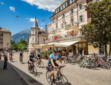 Group of cyclists riding through a cobblestone square in Merano, passing historic buildings, outdoor cafés, and rows of parked bicycles. Mountains rise in the background under a sunny blue sky.
