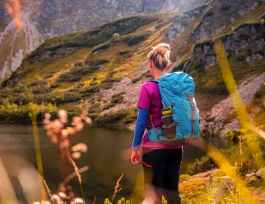 Hiker with a blue backpack stands beside a small alpine lake, looking across a rugged mountain landscape illuminated by warm sunlight. Rocky slopes and alpine vegetation surround the water beneath a dramatic mountain backdrop.