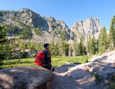 Hiker with a red backpack resting on a boulder in a mountain valley, surrounded by evergreen trees and rugged granite peaks under a clear blue sky.