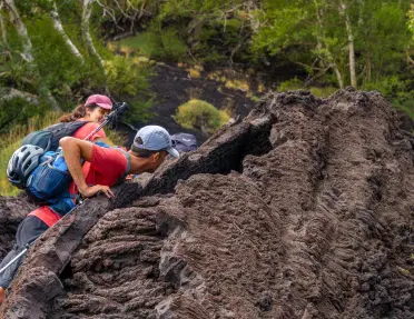 Man and woman climbing a large, dirt hill with tall trees in the background