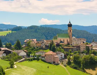 Panoramic view of a village nestled among rolling green hills and forests, centered around a historic church with an onion-domed bell tower and surrounded by traditional alpine buildings.