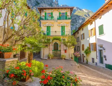 Charming village courtyard featuring colorful flowers, potted plants, and a historic building with green shutters and balconies. Steep rocky mountains rise behind the village beneath a clear blue sky.