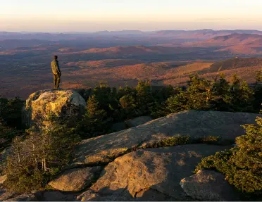 A hiker stands on a rock overlooking the sunlit mountain range in Vermont.