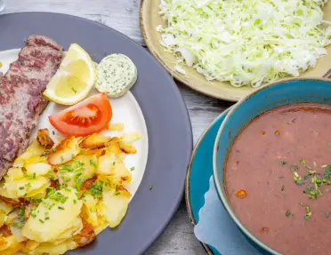 A meal featuring a grilled steak served with bratkartoffeln (German-style pan-fried potatoes), a cabbage salad and a bowl of soup.