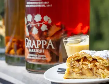 Slice of apple strudel dusted with powdered sugar served on a plate beside a small dessert and bottles of grappa, with the labels and bottles softly blurred in the background.