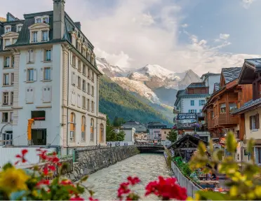 Exterior view of large white building next to a river, with snowy mountains in the distance