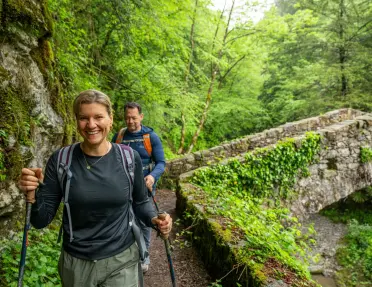 A hiker smiling hiker and her friend hike across a cobblestone bridge in a lush Italian forest.