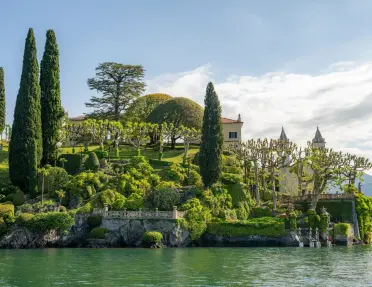 A beautiful collection of trees and other plants surround a building that sits on the shore of Lake Como.