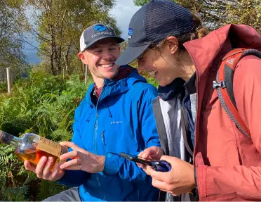 Two men smiling while hiking on a trail, looking at a bottle of liquor