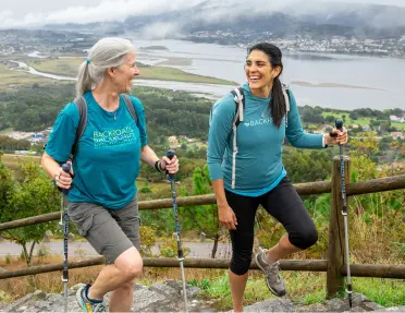 Two women are smiling and laughing together while hiking up a stone staircase outdoors on a cloudy day.
