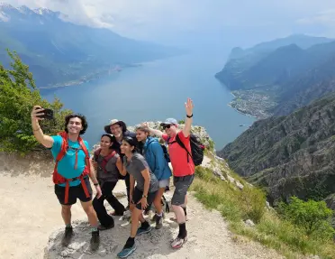 Group of hikers pauses to take a photo at a mountain viewpoint overlooking a large lake nestled between steep mountain slopes. The panoramic landscape stretches into the distance under hazy skies.