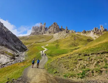 Hikers walk along a winding trail through green alpine hills toward dramatic jagged rock spires rising above the landscape. The mountain scenery is bathed in bright sunlight beneath a cloudless sky.