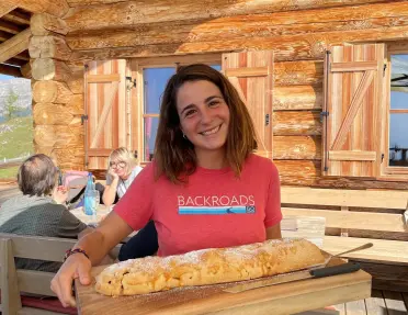 Person standing on the terrace of a rustic wooden mountain lodge, presenting a large powdered-sugar strudel on a wooden serving board. The alpine-style cabin features log walls, wooden shutters, and outdoor seating.