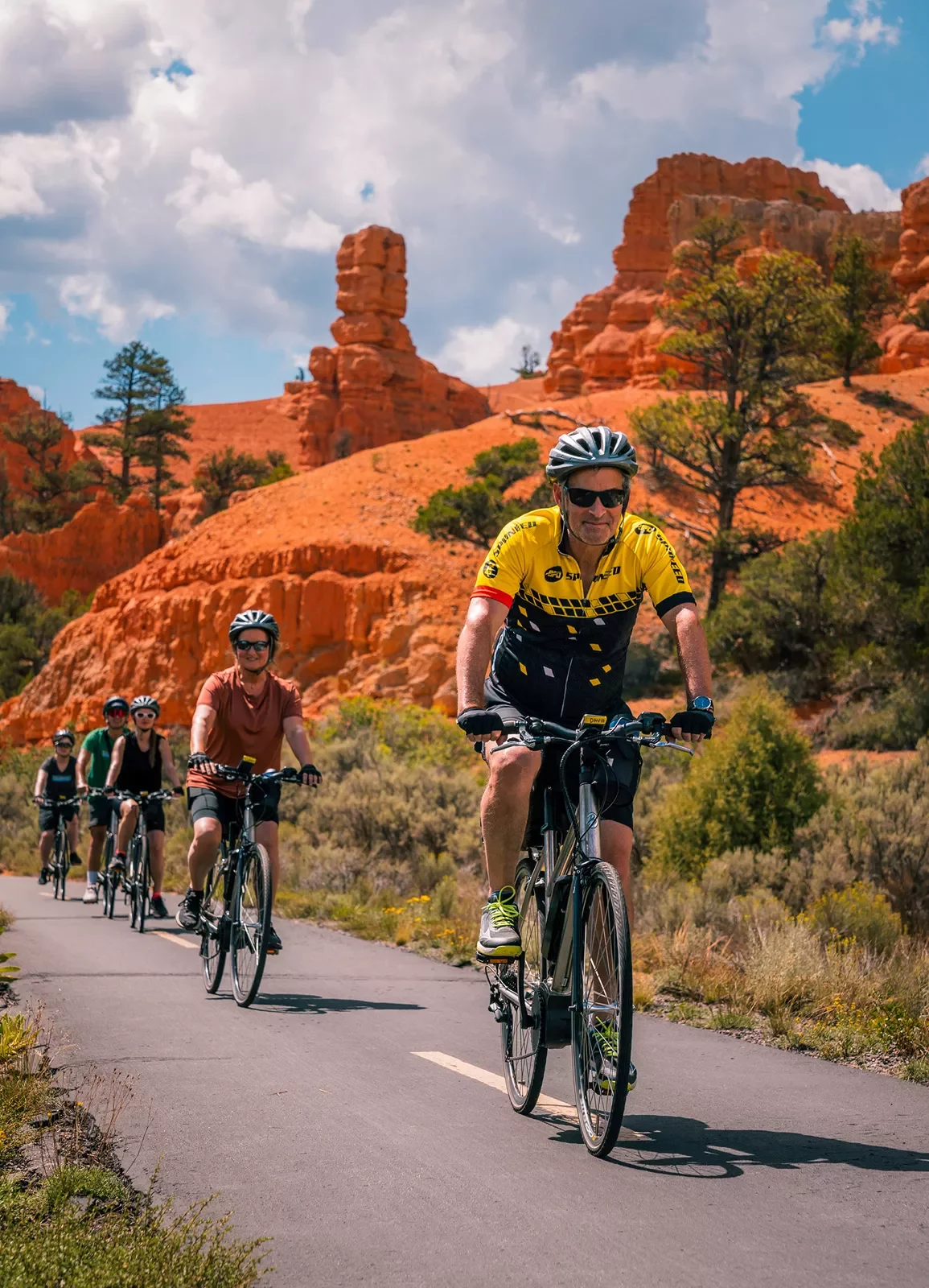 Five guests cycling towards camera, trees, vibrant red sand, hoodoos behind them.