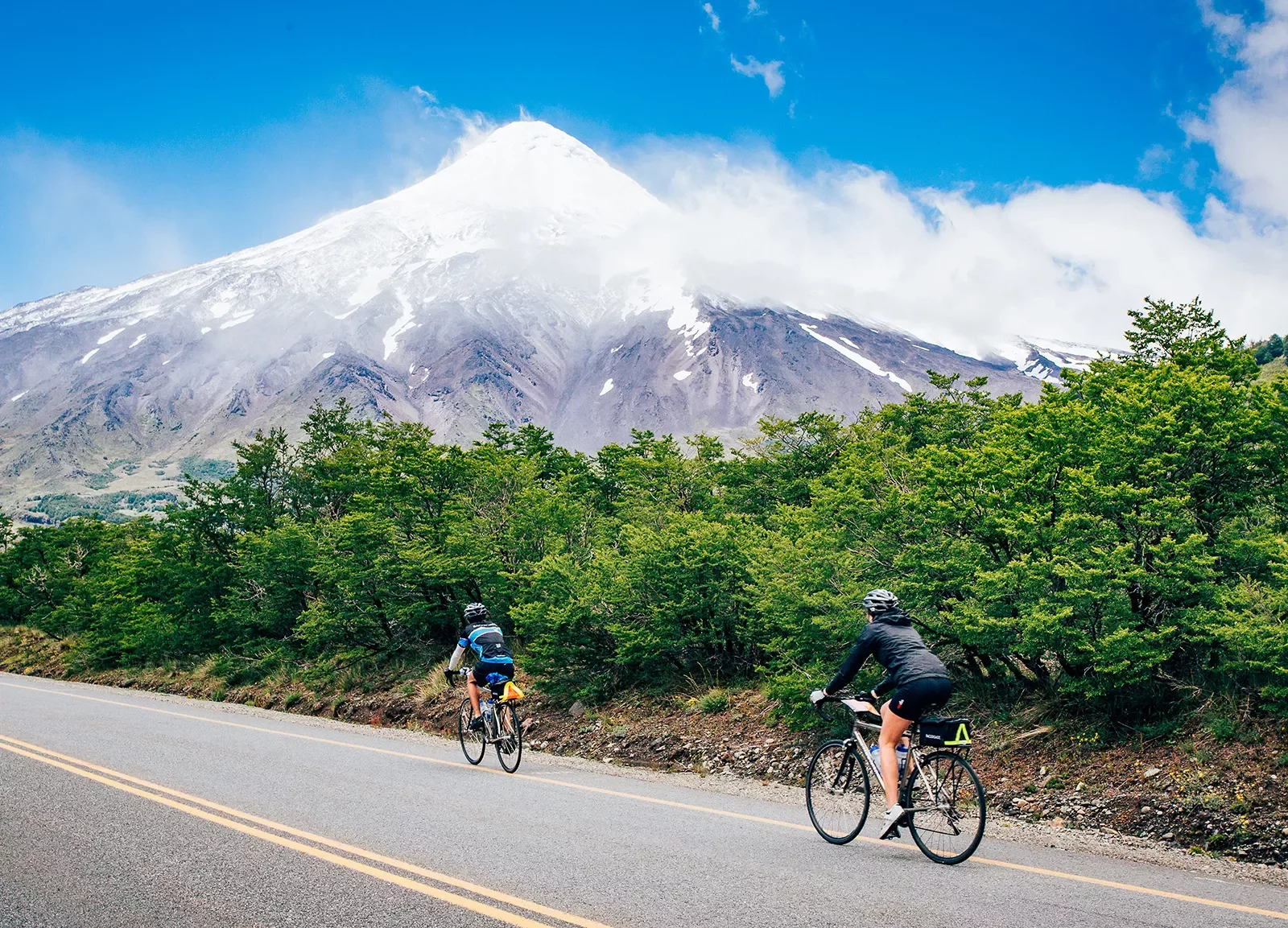 Two guests cycling up road, forest, snowy mountain in distance.
