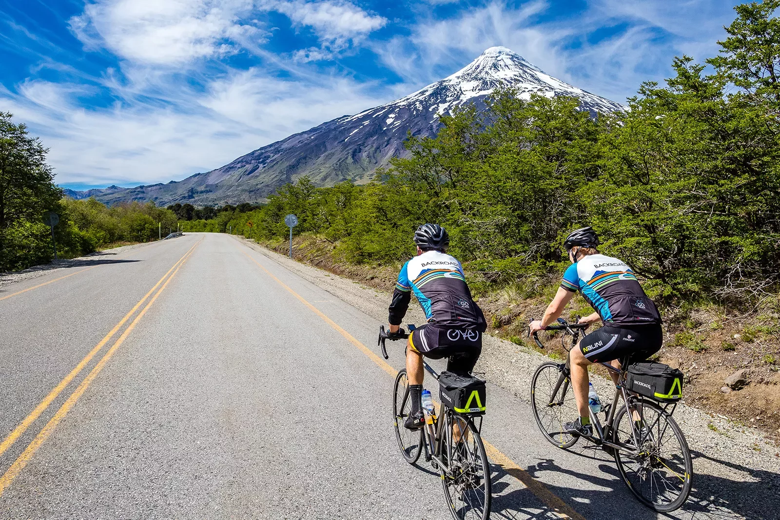 Two guests cycling down mountain road, snowy peak in background.