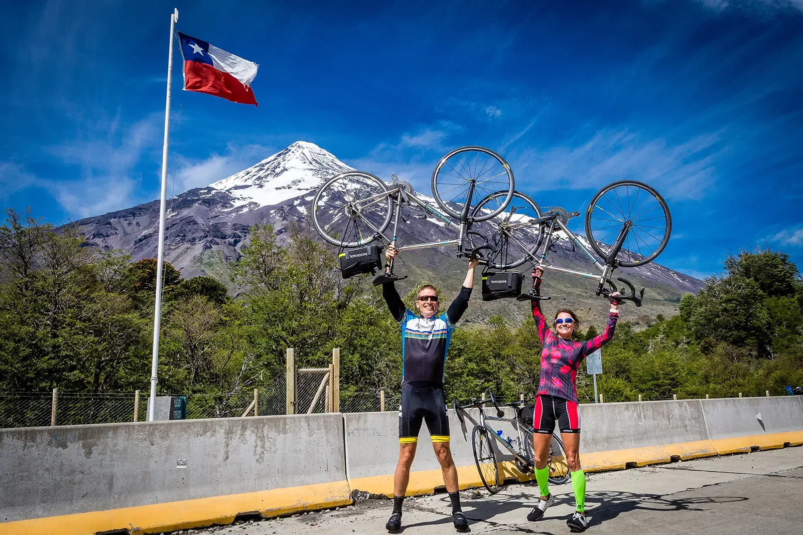 Two guests on road, holding bikes over head, Chilean flag, mountain behind them.