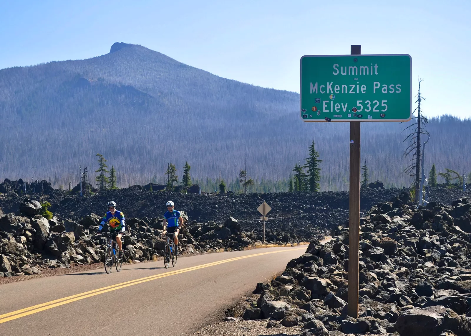 Two guests cycling past &quot;MCKENZIE PASS&quot; sign.