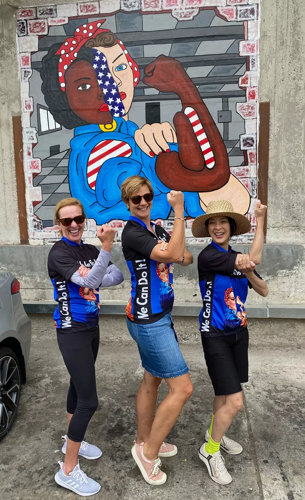 Three guests posing in front of Rosie the Riveter mural, doing her pose.