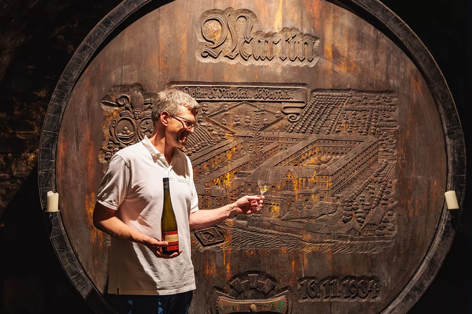 Man holding a bottle of wine in one hand and a glass with wine in the other in front of a very large wine barrel