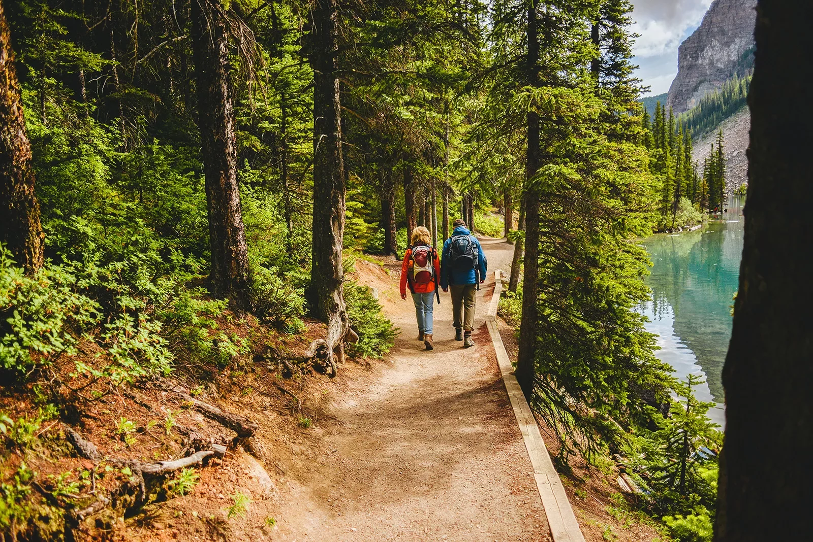 Two guests hiking down forest trail, river or lake to their right.