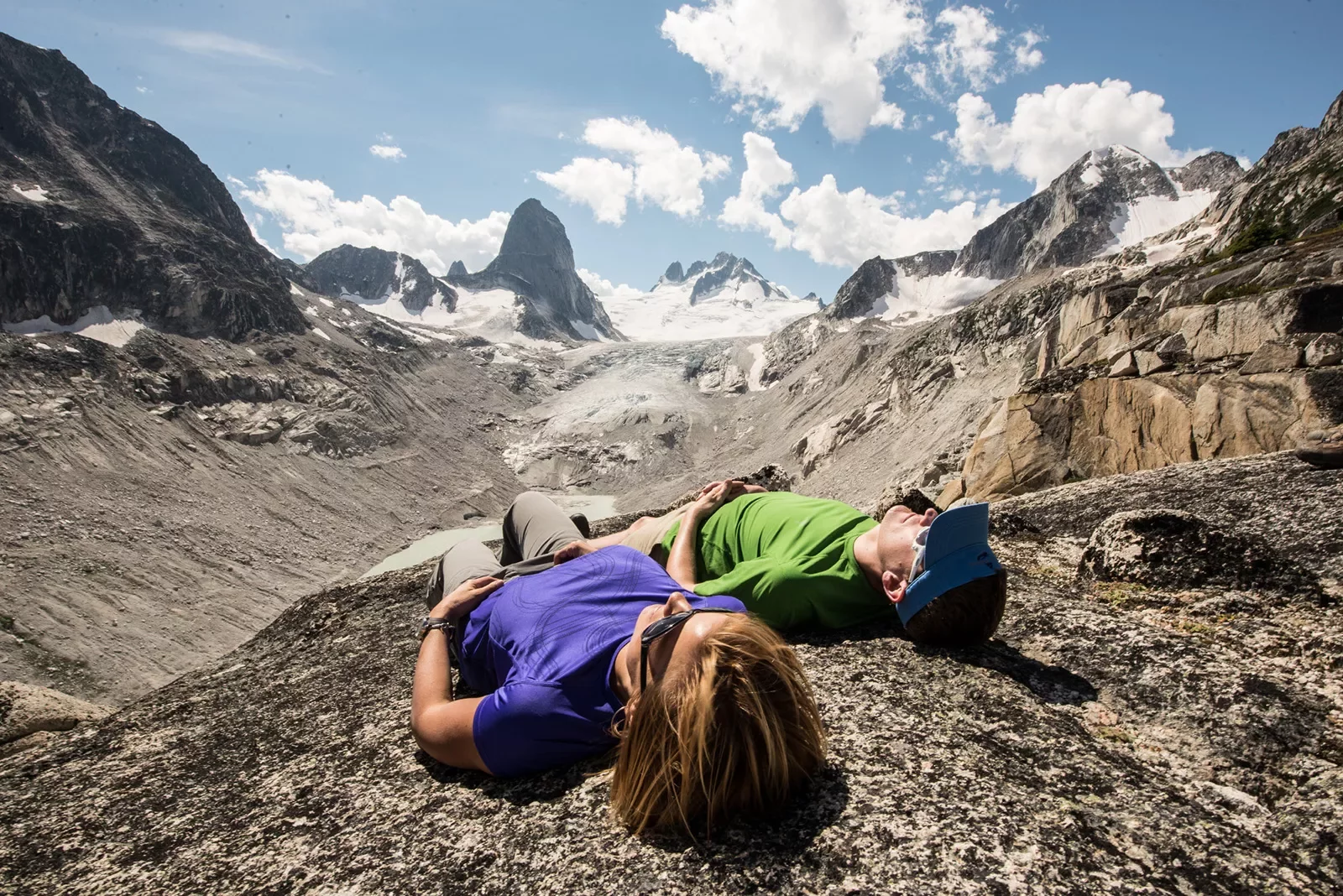 Two guests laying on rocky mountainside, arid cliffs and clouds in background.
