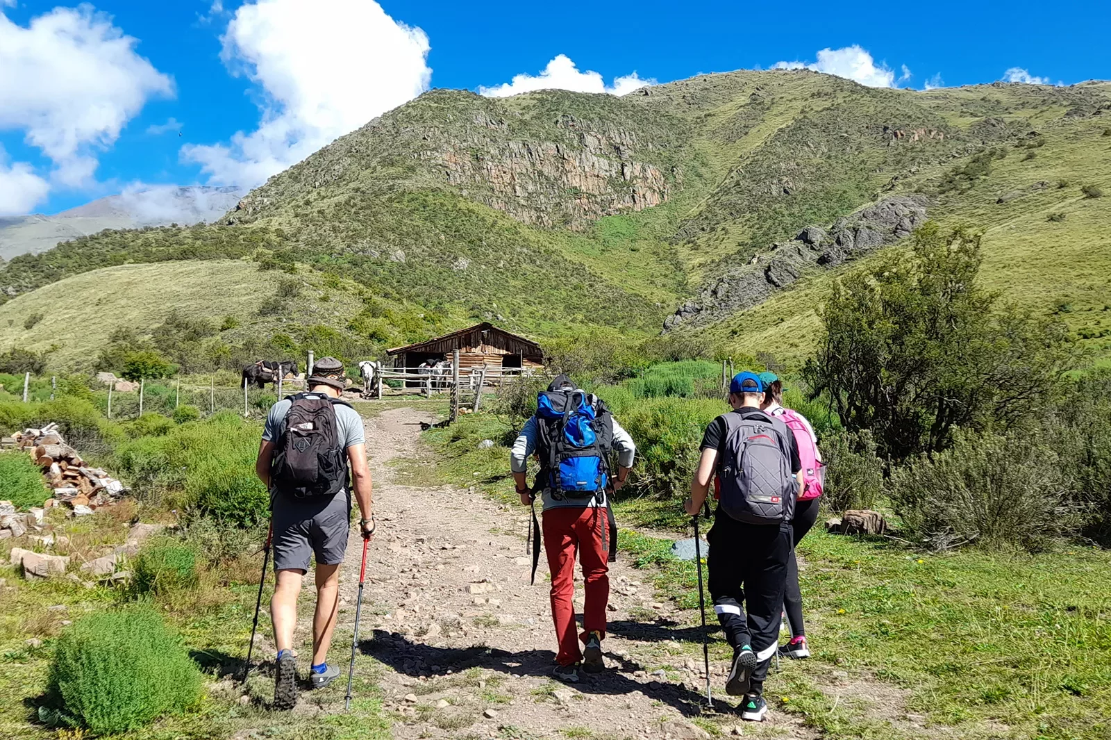 Three guests hiking up to a wooden farm house. Mountains in distance.