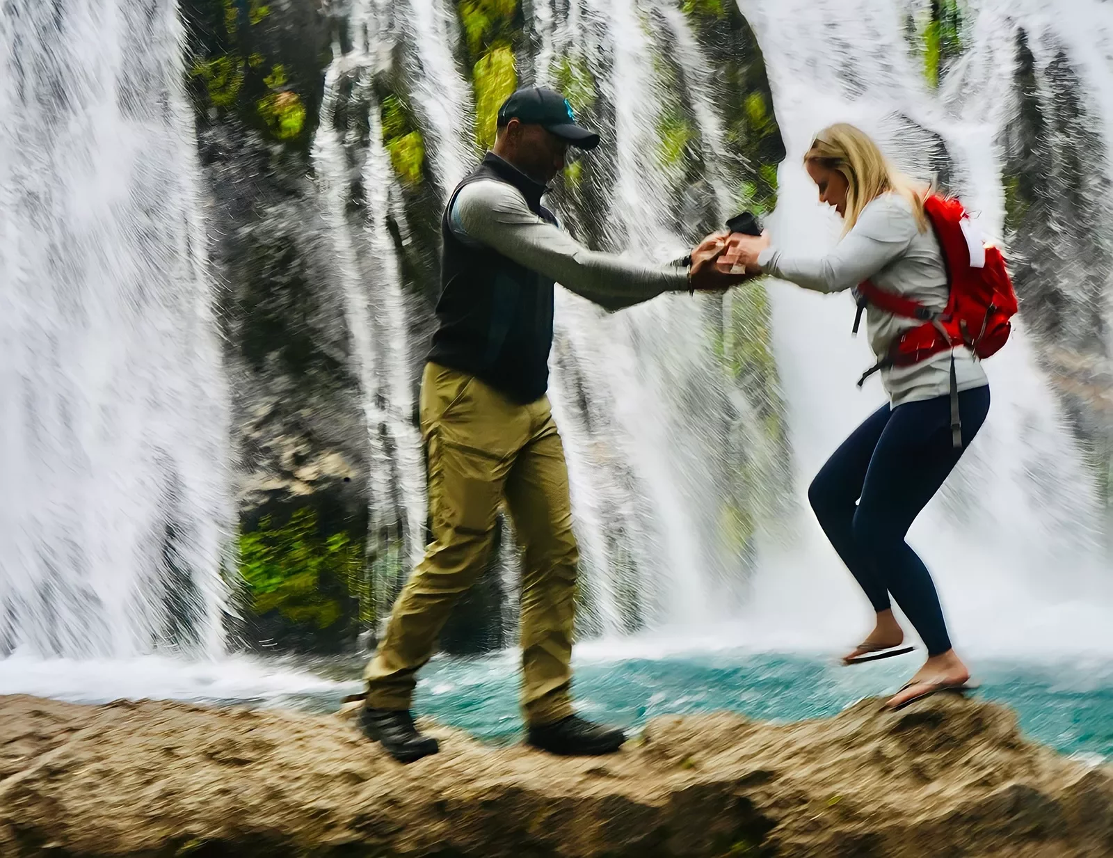 Two guests walking over rocks, waterfall behind them.