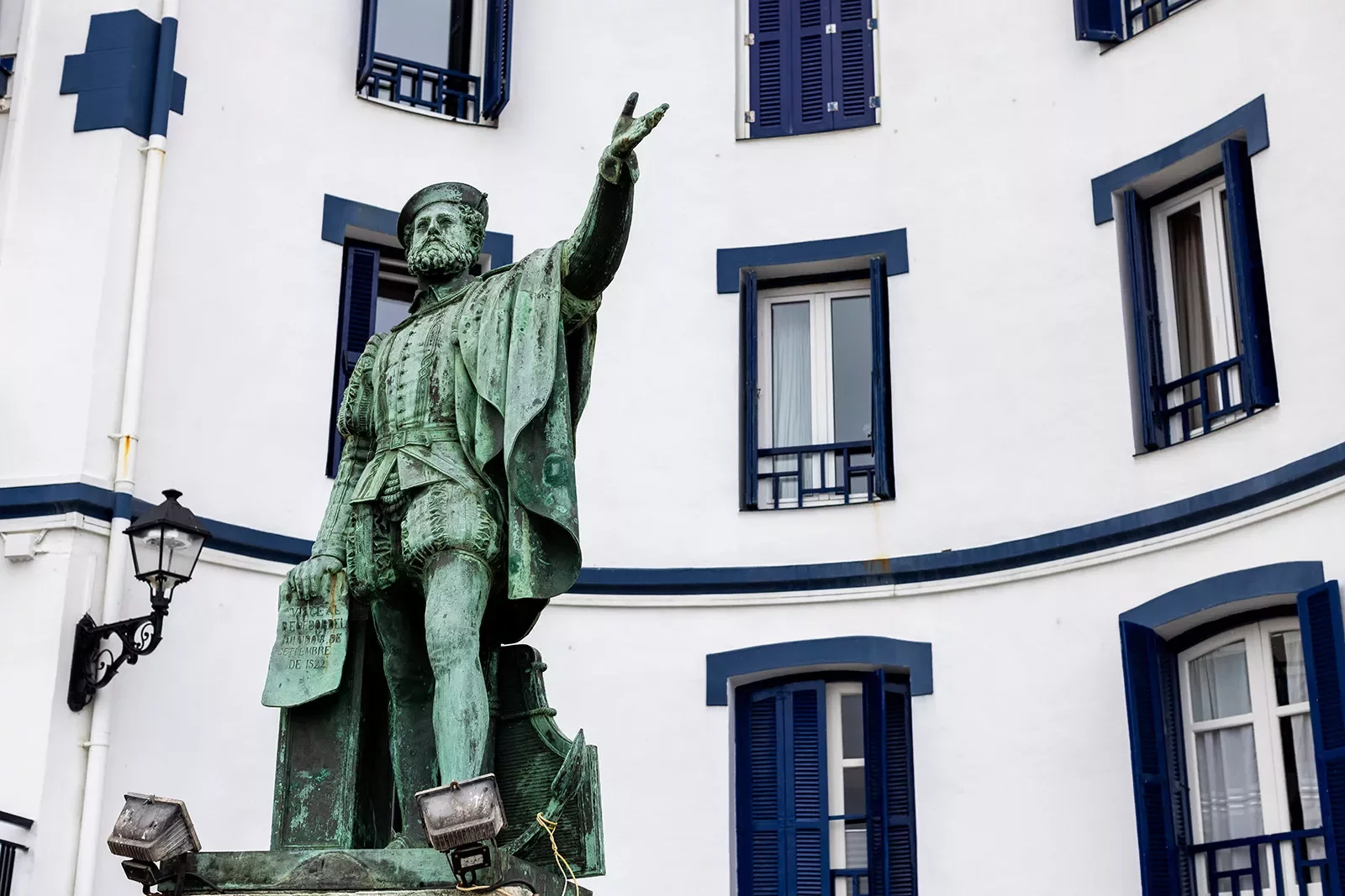 Patinaed copper statue of Juan Sebastian Elcano, blue and white building behind.