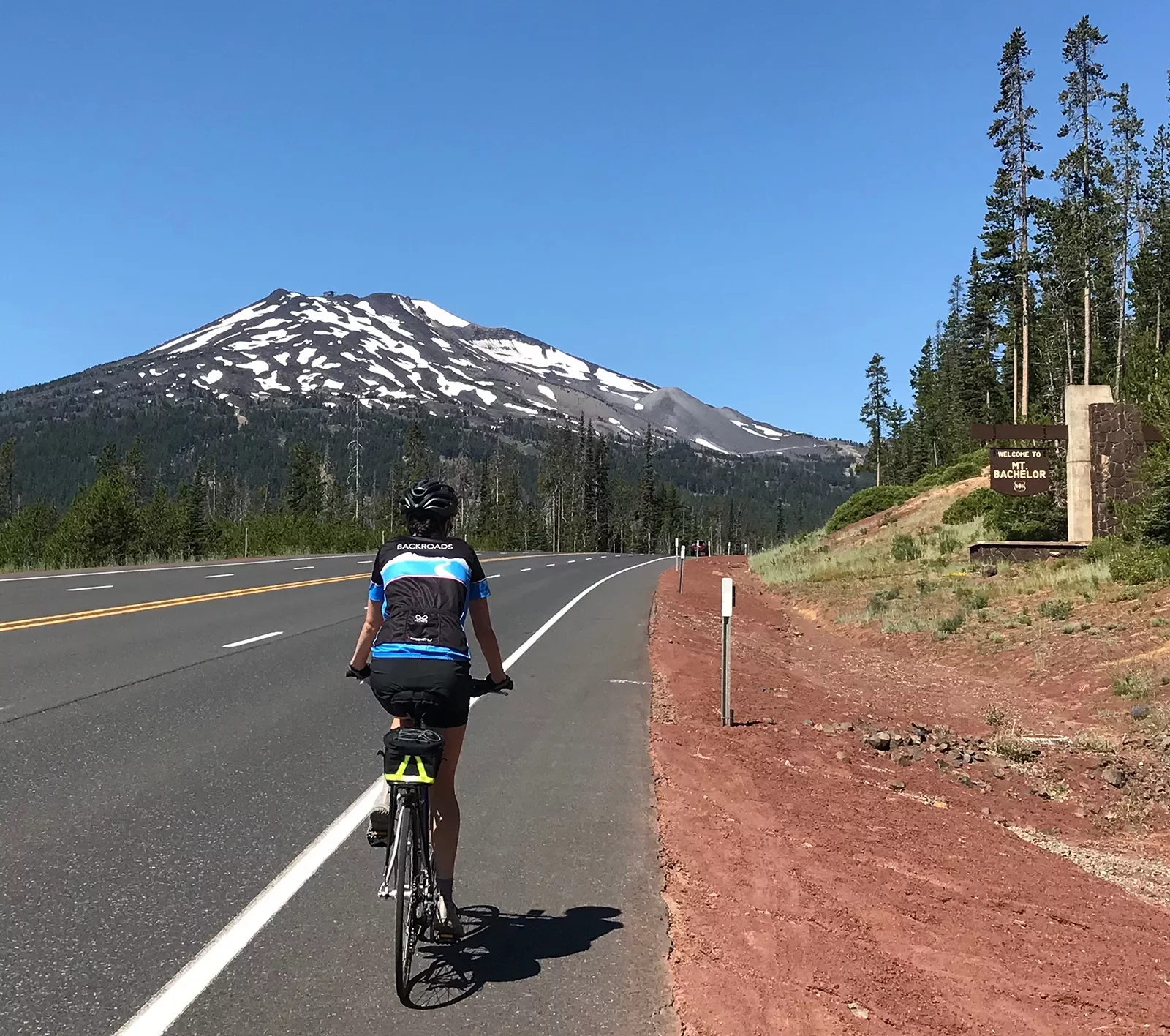 Guest cycling past &quot;MT. BACHELOR&quot; sign, the mountain itself in the background.