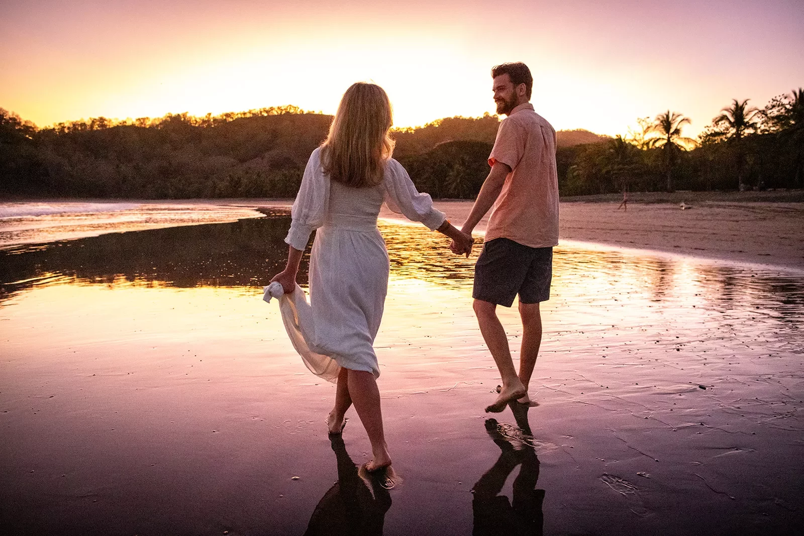 Two guests holding hands, walking along shore during sunset.